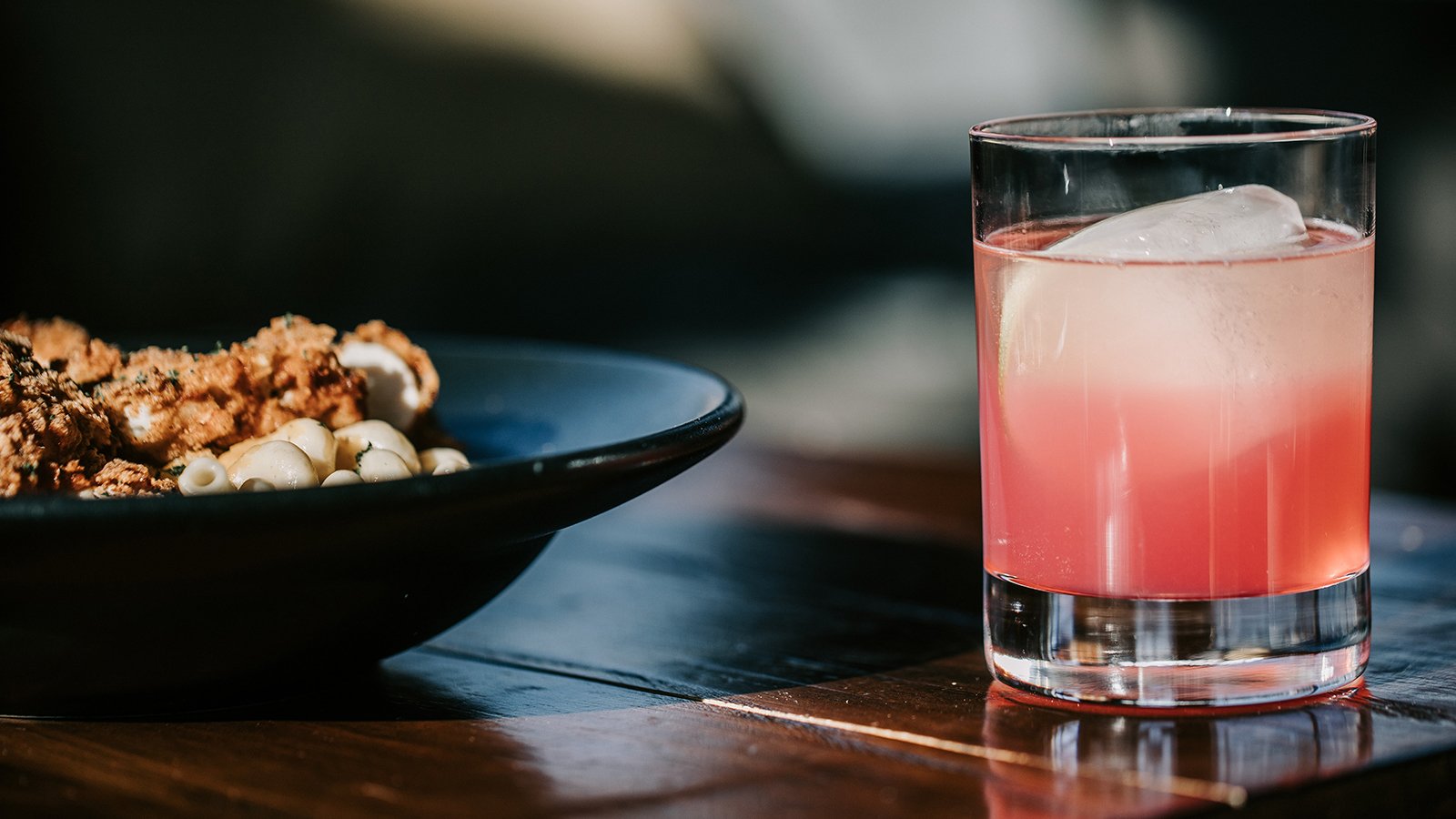 A closeup of a pink cocktail and a plate of food on a wooden table.