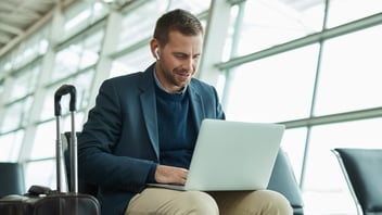 Business traveler in suit works on laptop with wireless earbuds in bright modern airport terminal.