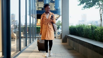 Business traveler in tan coat checks phone while pulling luggage through modern urban walkway with greenery.