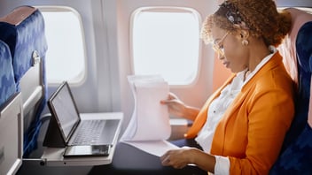 A businesswoman sitting in an airplane using a laptop and reviewing printed documents.