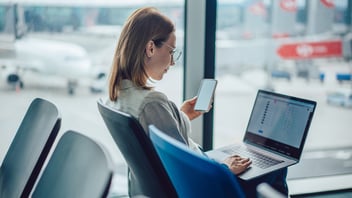 Young female business traveler using a smartphone and laptop while waiting for her flight at an airport.