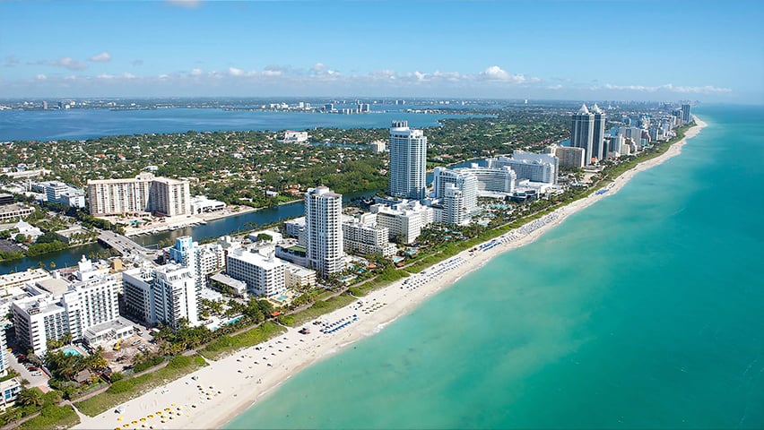Aerial view of Miami Beach showing white sand beaches, turquoise ocean waters, and high-rise hotels and condominiums lining the coastline.