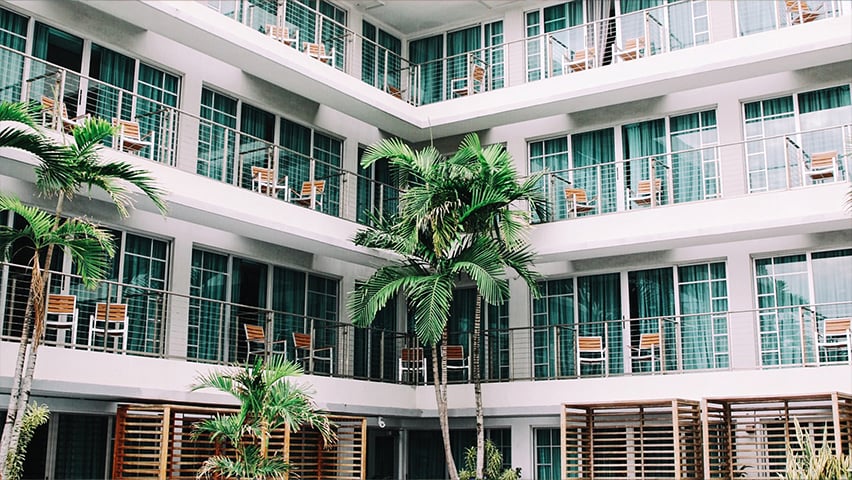 Modern white hotel or resort building with multiple floors, glass balconies, and palm trees in a central courtyard.