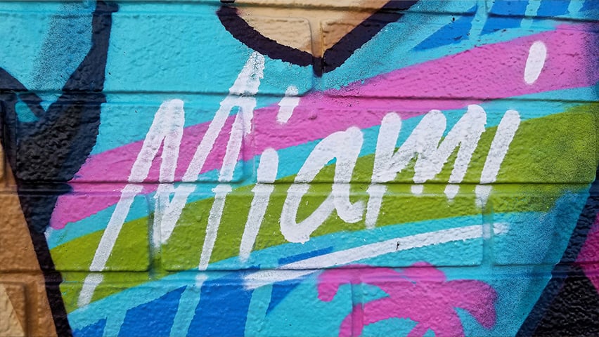 Colorful street art mural featuring "Miami" in white letters against vibrant blue, pink, and green geometric patterns.