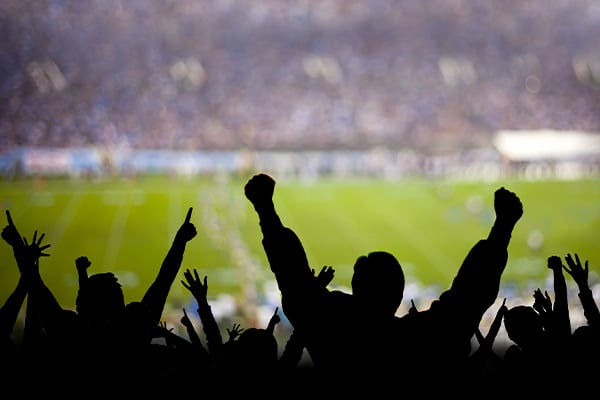 Silhouetted crowd of sports fans cheering with raised arms at a stadium with bright field lights in background.