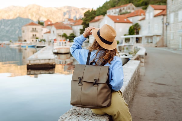 Tourist with backpack and hat sits by waterfront overlooking Mediterranean coastal town with boats and buildings.