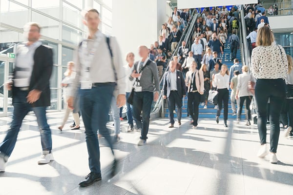 Blurred motion of busy attendees walking and using escalators at a trade show in a modern convention center.