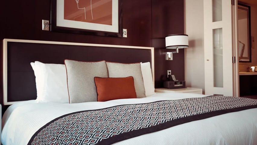 Modern hotel room with white bedding, geometric throw blanket, and orange accent pillow.