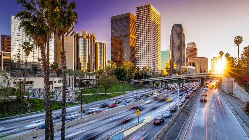 Downtown Los Angeles skyscrapers at golden hour with busy freeway traffic streaks in the foreground.