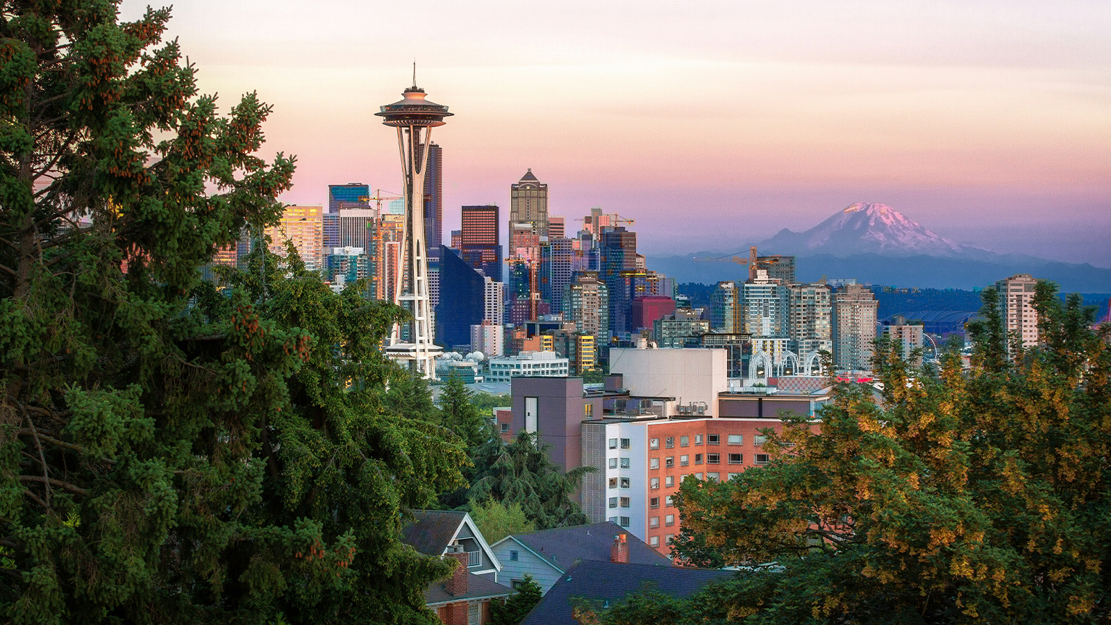 The Seattle skyline at sunset with Mount Rainier in the background and evergreen trees in the foreground.