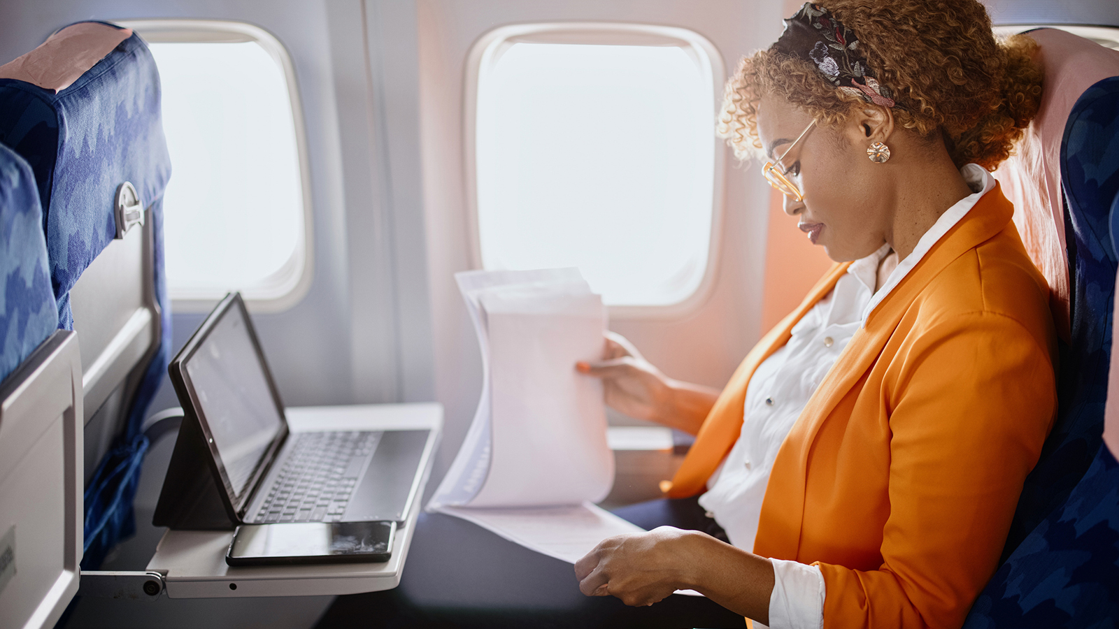 A businesswoman sitting in an airplane using a laptop and reviewing printed documents.