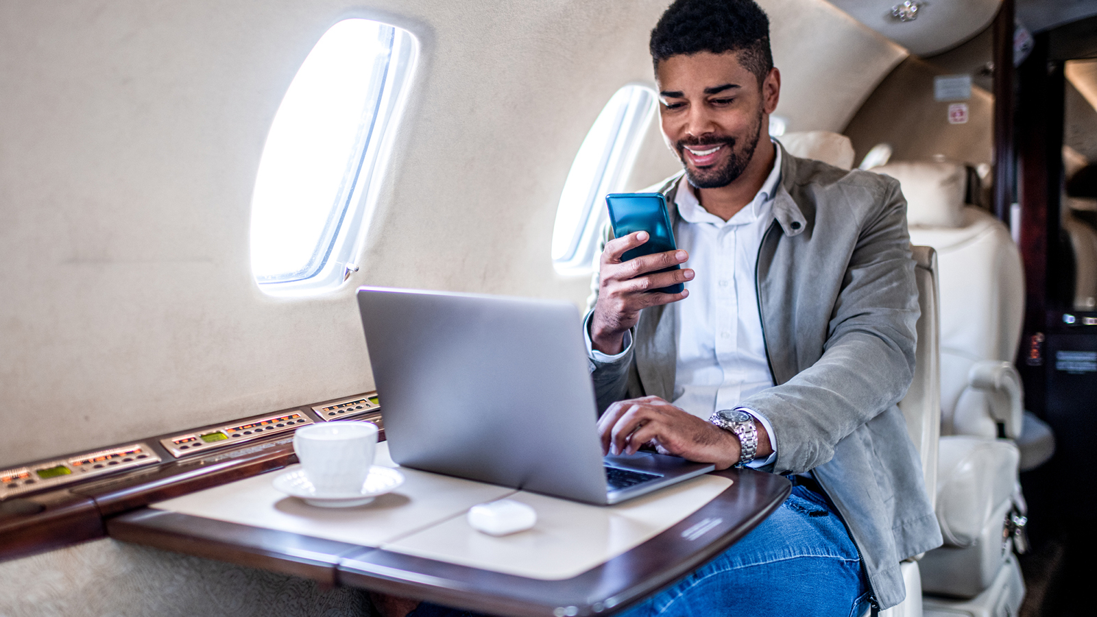 Young casually dressed businessman smiles while looking at his smart phone during the first class flight in an airplane.