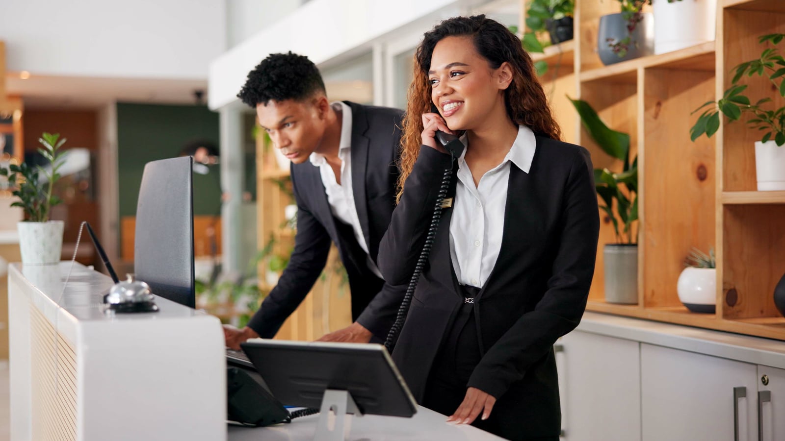Two hotel front desk staff in black blazers assist guests, with one smiling while speaking on a phone and the other working at a computer.